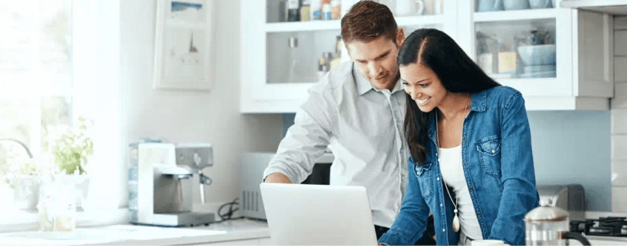 A smiling couple in the kitchen looks at something on their laptop